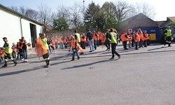 Eine Gruppe von Menschen in orangefarbenen Warnwesten geht und reinigt die Straße in einem Außenbereich. Bäume und ein Gebäude sind hinter ihnen.