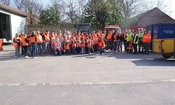 Eine Gruppe von Menschen in orangefarbenen Westen posiert für ein Foto vor einem Gebäude. Hinter ihnen stehen einige Maschinenfahrzeuge, Bäume und ein klarer Himmel.