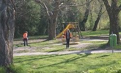 Zwei Personen gehen in einem Park mit einem gelben Spielgerät und einem grünen Mülleimer. Bäume und Gras umgeben den Bereich.