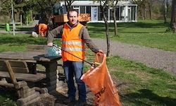 Ein Mann in einer reflektierenden orangefarbenen Weste hält einen Müllsack in der Hand, während er neben einer Picknick-Bank im Park steht.