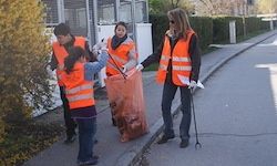 Eine Gruppe von Menschen in Sicherheitswesten sammelt Müll am Straßenrand. Eine Person hält einen Besen, während eine andere eine Tasche hält.