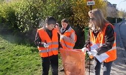 Drei Personen in reflektierenden orangefarbenen Westen sammeln Müll in der Nähe einer Straße, mit einem Schild und Grün im Hintergrund.