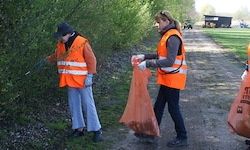 Zwei Frauen in orange Sicherheitswesten und Handschuhen sammeln Müll entlang eines unbefestigten Weges in der Nähe von Grünflächen. Eine hält einen Müllsack, während die andere neben einem Busch steht.