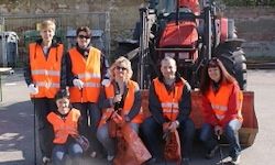 Eine Gruppe von Menschen in orange Sicherheitswesten posiert vor einem roten Traktor. Einige halten Taschen und einer trägt Handschuhe. Im Hintergrund befinden sich eine Steinmauer und ein Metallzaun.