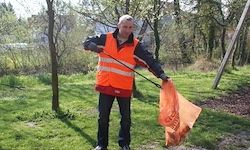 Ein Mann in einer reflektierenden Weste hält einen Plastikbeutel und sammelt Müll in einem Park mit Bäumen und Grün im Hintergrund.