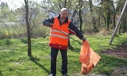 Ein Mann in einem orangefarbenen Sicherheitsweste steht in einem Grasbereich und hält einen Müllsack. Bäume und ein Holzpfahl sind im Hintergrund.