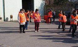 Eine Gruppe von Menschen in orangenen Westen steht in einer Reihe, möglicherweise auf einem Parkplatz. Sie scheinen Freiwillige zu sein, und einige halten Stöcke. Im Hintergrund befindet sich ein Gebäude und Bäume.