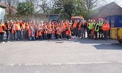 Eine Gruppe von Menschen in orangefarbenen Westen steht draußen. Hinter ihnen sind mehrere Fahrzeuge geparkt, darunter ein gelber Lastwagen. Bäume und Gebäude sind im Hintergrund zu sehen.