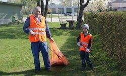 Ein erwachsener Mann und ein Kind sind in einem Park und tragen beide orange Westen. Sie halten einen Plastiksack und einen Stock und sammeln wahrscheinlich Müll.