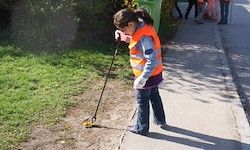 Ein junges Mädchen in einer reflektierenden Weste benutzt einen Metalldetektor auf dem Bürgersteig. Sie schaut nach unten auf den Boden, während sie den Detektor hält.
