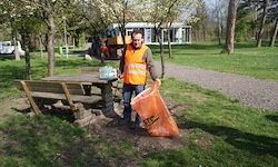 Ein Mann in einer reflektierenden orangefarbenen Weste steht neben einer Parkbank und hält einen großen orangefarbenen Müllsack. Der Park ist von Bäumen und Gras umgeben. Im Hintergrund ist ein Baufahrzeug zu sehen.