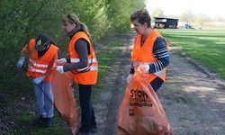 Drei Frauen in orangefarbenen Westen sammeln Müll entlang eines Feldwegs. Eine hält einen Plastikbeutel, eine andere einen Stock und die dritte hat Handschuhe und einen Beutel. Der Hintergrund zeigt Grünflächen und ein Feld.