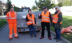 Vier Personen in reflektierenden orangefarbenen Westen stehen auf einer Straße, mit einem Lastwagen und Müllsäcken hinter ihnen.