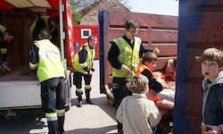 Rettungskräfte in gelben Warnwesten laden Ausrüstung in einen Lastwagen. Kinder beobachten von einer blauen Barriere aus. Die Szene spielt im Freien.