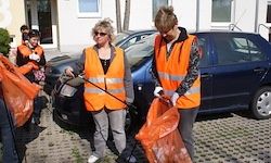 Zwei Frauen in orange Weste putzen ein blaues Auto, während andere in der Nähe sind. Eine hält ein Reinigungswerkzeug und die andere einen Beutel. Dahinter stehen ein paar Leute und ein Gebäude.
