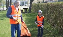 Ein Erwachsener und ein Kind in orangefarbenen Westen sammeln Müll in einem Park. Eine Bank und ein Busch sind im Hintergrund.