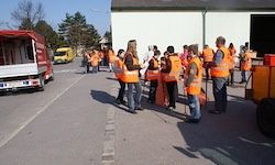Eine Gruppe von Freiwilligen in orange Westen steht auf der Straße, mit einem Gebäude, Fahrzeugen und Bäumen im Hintergrund.