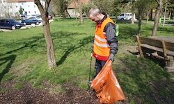 Ein Mann in einer orangefarbenen Weste sammelt Müll in einem Park. Hinter ihm steht ein Baum, eine Bank und mehrere geparkte Autos.