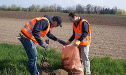 Zwei Männer in orange Weste und Handschuhen säubern ein Feld von Müll, einer hält einen Beutel.