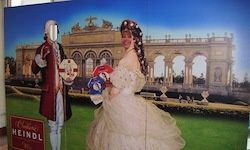 Eine Ausschnitt von einem Mann und einer Frau, gekleidet in historischer Kleidung, steht vor einer Kulisse eines historischen Gebäudes. Die Frau hält einen Blumenstrauß.