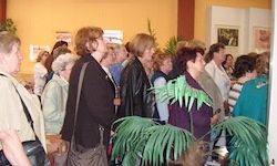 Eine Gruppe von Menschen steht in einem Raum mit gelben Wänden, Pflanzen und Bilderrahmen. Einige tragen Jacken, andere haben Taschen. Sie scheinen miteinander zu sprechen.