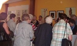 Eine Gruppe älterer Menschen steht und unterhält sich in einem Raum mit gelben Wänden, einer weißen Decke und gerahmten Kunst an den Wänden.