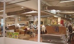 Ein Fabrikinnenraum mit Arbeitern in weißen Kitteln und Schutzhelmen, die Maschinen bedienen. Ein Glasfenster zeigt eine Reflexion der Fabrik. Stapel von Kartons und Lagerboxen sind sichtbar.