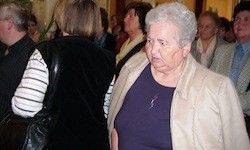 Eine ältere Frau mit weißen Haaren steht unter einer Gruppe von Menschen und wirkt nachdenklich, während sie vorbeigeht.