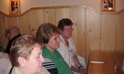 Vier Frauen sitzen in einer hölzernen Nische und schauen auf etwas vor sich. Hinter ihnen befinden sich Holzwände und Bilderrahmen.