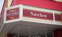 Ein Laden mit einem roten und weißen Vordach, das 'Heindl Naschen' in fett roten Buchstaben auf weißem Hintergrund zeigt. Das Schild hat ein Vintage-Aussehen mit rotem Rand.