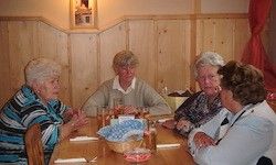 Fünf ältere Frauen sitzen um einen Holztisch und unterhalten sich. Auf dem Tisch befinden sich ein Korb mit Brot, ein Glas Bier und eine Flasche Wein. Hinter ihnen ist eine Holzwand.