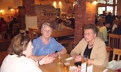 Drei Frauen sitzen an einem Tisch in einem Restaurant und unterhalten sich. Der Tisch ist mit Gläsern und einer Speisekarte besetzt. Hinter ihnen sitzen andere Gäste an Tischen und in der Nähe eines Kamins.