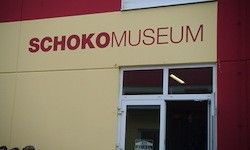 Ein gelbes Gebäude mit rotem Rand hat ein Schild mit der Aufschrift 'SCHOKO MUSEUM' über der Eingangstür. Die Tür ist weiß mit einem Glasfenster und ist leicht geöffnet.