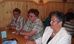 Vier Frauen sitzen an einem Holztisch, zwei tragen eine Brille. Die Frau in Weiß schaut auf etwas auf dem Tisch, während eine andere Frau in einem karierten Shirt gestikuliert. Hinter ihnen sitzt eine weitere Frau in einem grünen Shirt.