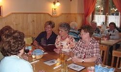 Eine Gruppe von Menschen sitzt in einem Restaurant an einem Tisch und plaudert, während sie ihr Essen genießt. Der Tisch ist mit Tellern, Gläsern und Servietten gedeckt. Eine Frau liest eine Speisekarte.