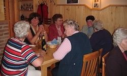 Eine Gruppe älterer Frauen sitzt um einen Holztisch und isst. Einige von ihnen unterhalten sich und lachen, während andere etwas auf dem Tisch betrachten.