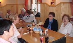 Mehrere ältere Frauen sitzen um einen Holztisch. Sie unterhalten sich. Flaschen und Gläser stehen auf dem Tisch. Eine Pflanze steht in der Ecke.