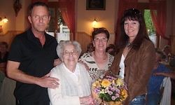 Vier Erwachsene, darunter eine ältere Frau mit Brille, stehen zusammen. Ein Mann und eine Frau halten Blumen. Sie sind bei einer Feier, wahrscheinlich einem Geburtstag.