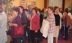 Eine Gruppe von Menschen steht in einer Reihe, einige unterhalten sich und andere sehen sich um, tragen verschiedene Kleidungsstücke und Accessoires, mit einer Pflanze und Rahmen an der Wand.