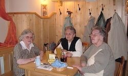 Drei ältere Frauen sitzen an einem Holztisch mit Getränken und Essen in einem Raum mit Holzwänden und aufgehängten Mänteln.