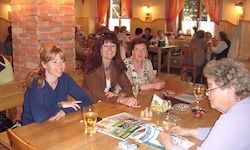 Vier Frauen sitzen an einem Tisch in einem Restaurant. Zwei haben Weinglas, eine hat ein Bier. Eine Frau trägt eine Brille. Eine Speisekarte, Salz und Pfefferstreuer sind auf dem Tisch.