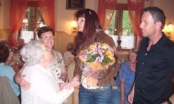 Eine ältere Frau in einem weißen Kleid hält die Hände einer Frau mit Blumen, die neben einem Mann in einem schwarzen Hemd steht, in einem Raum mit roten Vorhängen und Lampen.