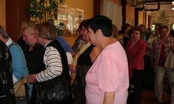 Eine Gruppe von Menschen steht in einem Raum und unterhält sich. Eine Frau in einem rosa Shirt steht vorne in der Mitte und hält eine Handtasche.