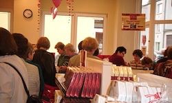 Eine Gruppe von Menschen steht in einer Schlange in einem Geschäft. Sie sind von Regalen umgeben, die mit verschiedenen Produkten gefüllt sind, möglicherweise Schokolade. Eine Uhr ist an der Wand montiert.