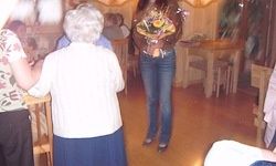 Eine Frau in Jeans steht mit einem Blumenstrauß in einem Raum und sieht eine ältere Frau in einem weißen Shirt an. Hinter ihnen sitzen Menschen um einen Tisch herum. Der Raum hat Holzböden und Stühle.
