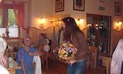 Eine Frau mit einem Blumenstrauß steht in einem Esszimmer mit einigen Personen, die an Tischen sitzen. Es gibt Lampen, Bilderrahmen und ein Fenster mit Vorhängen.