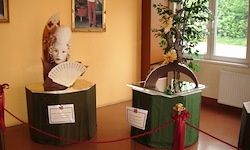 Zwei grüne Ständer mit Skulpturen, einer mit einer Maske und einem Fächer, der andere mit einem Baum, in einem Raum mit gelben Wänden und einem Fenster.