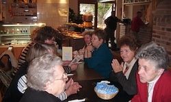 Eine Gruppe älterer Frauen sitzt in einem Restaurant um einen Tisch, klatscht in die Hände und lächelt. Ein Korb steht auf dem Tisch und einige Leute stehen im Hintergrund.