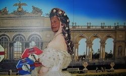 Eine Person in einem weißen Kleid steht vor einem großen, klassischen Gebäude mit mehreren Bogenfenstern und hält ein rotes und blaues Schild.