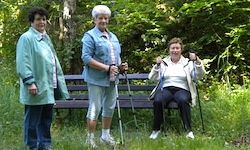 Drei ältere Frauen stehen draußen in einem Park mit einer Bank und Gehstöcken. Eine Frau sitzt auf der Bank und die anderen stehen.